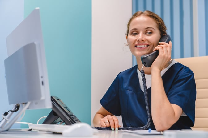 Smiling-young-woman-nurse-receptionist-talking-on-phone-while-working-in-modern-clinic-1275071451_7952x5304